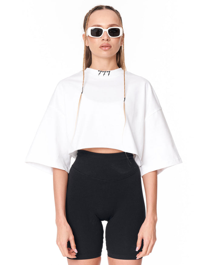 A model wearing a white cropped t-shirt with a high mock neck and wide short sleeves, paired with black bike shorts and white-framed sunglasses. The shirt features the number '777' printed in black near the collar.