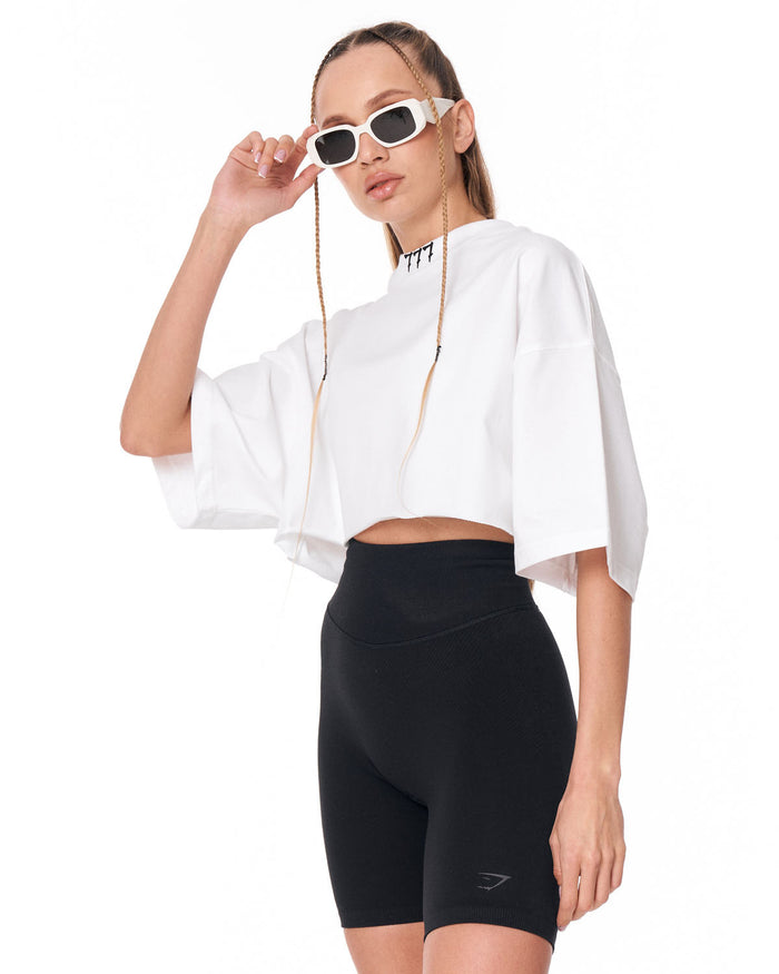 A model wearing a white cropped tunic top with wide sleeves, black high-waisted biker shorts, and white-framed sunglasses with chain straps, posed against a solid white background.