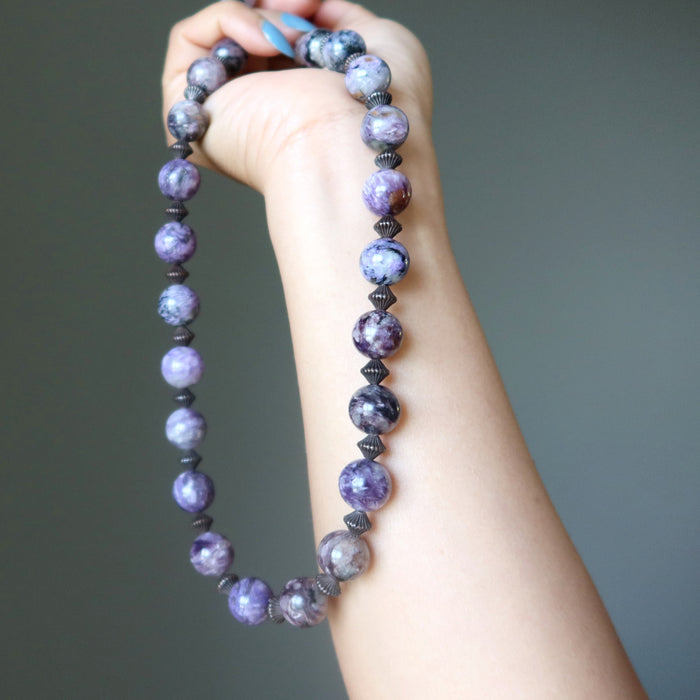 Hand holding a charoite beaded necklace with purple and black beads against a gray background
