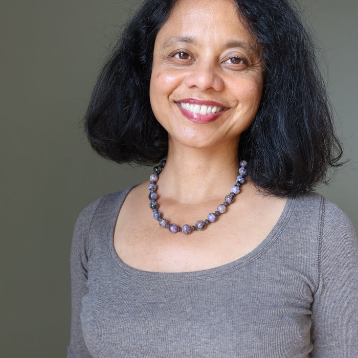 Woman wearing a gray shirt and purple charoite beaded necklace against a plain background