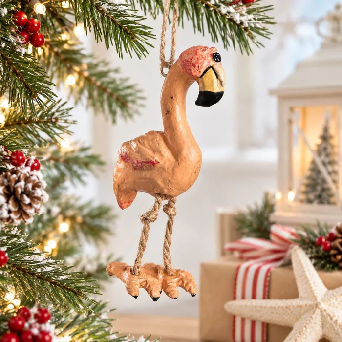 A rustic wooden flamingo Christmas ornament hanging from a pine tree branch with warm holiday lights, wrapped in a cozy festive setting with wrapped gifts and a lantern in the background.